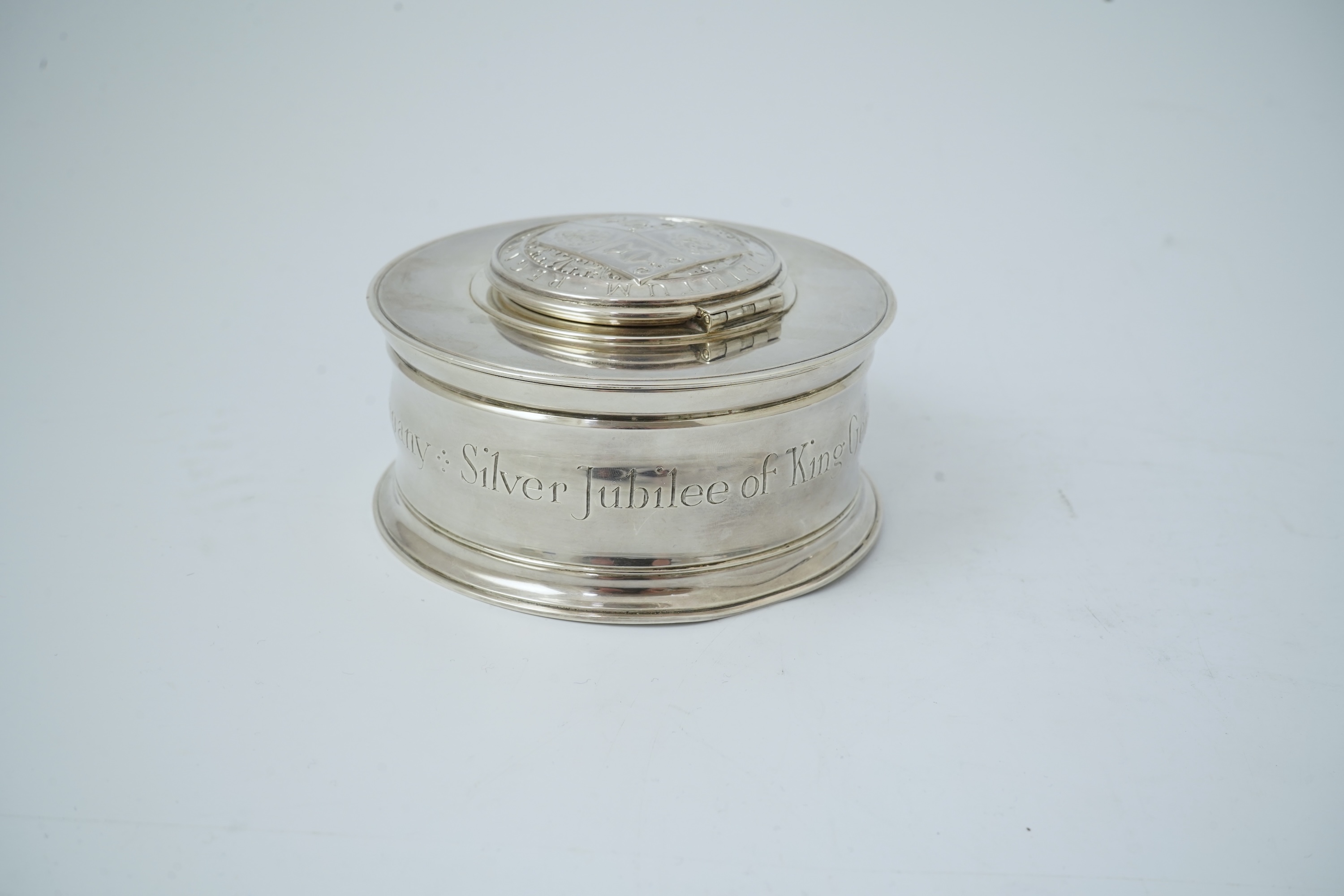 A George V silver circular inkwell, by Sebastian Garrard, commemorating the Silver Jubilee of King George V, the die stamped cover designed by George Kruger Gray featuring the Goldsmiths Company Coat of Arms